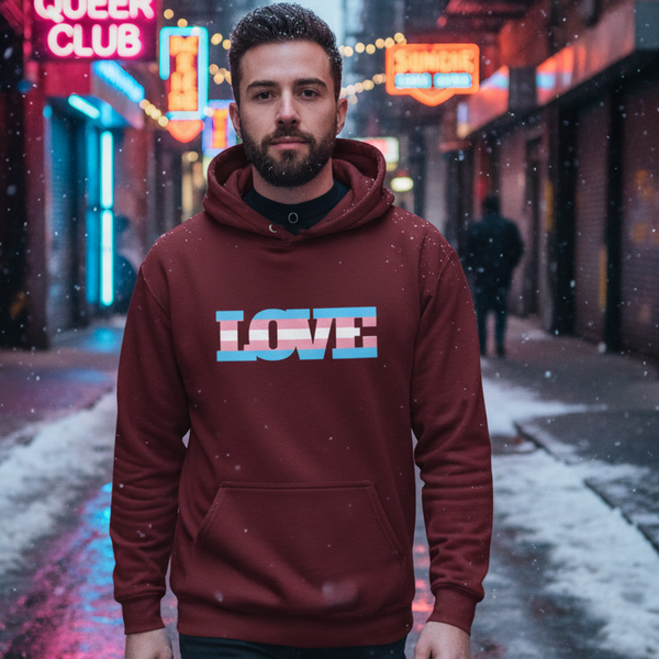 Bearded person wearing a burgundy hoodie with LOVE in transgender-pride stripes in a snowy neon-lit alley