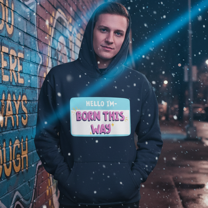 Person wearing a dark hoodie reading “HELLO I'M—BORN THIS WAY” beside a graffiti wall