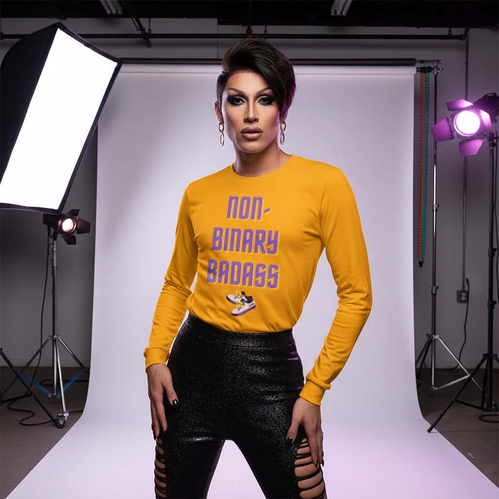 Non-Binary Badass Unisex Long Sleeve T-Shirt in mustard-yellow with bold purple lettering, worn by a non-binary model styled with dramatic makeup and glittery cut-out pants, exuding empowerment and pride.