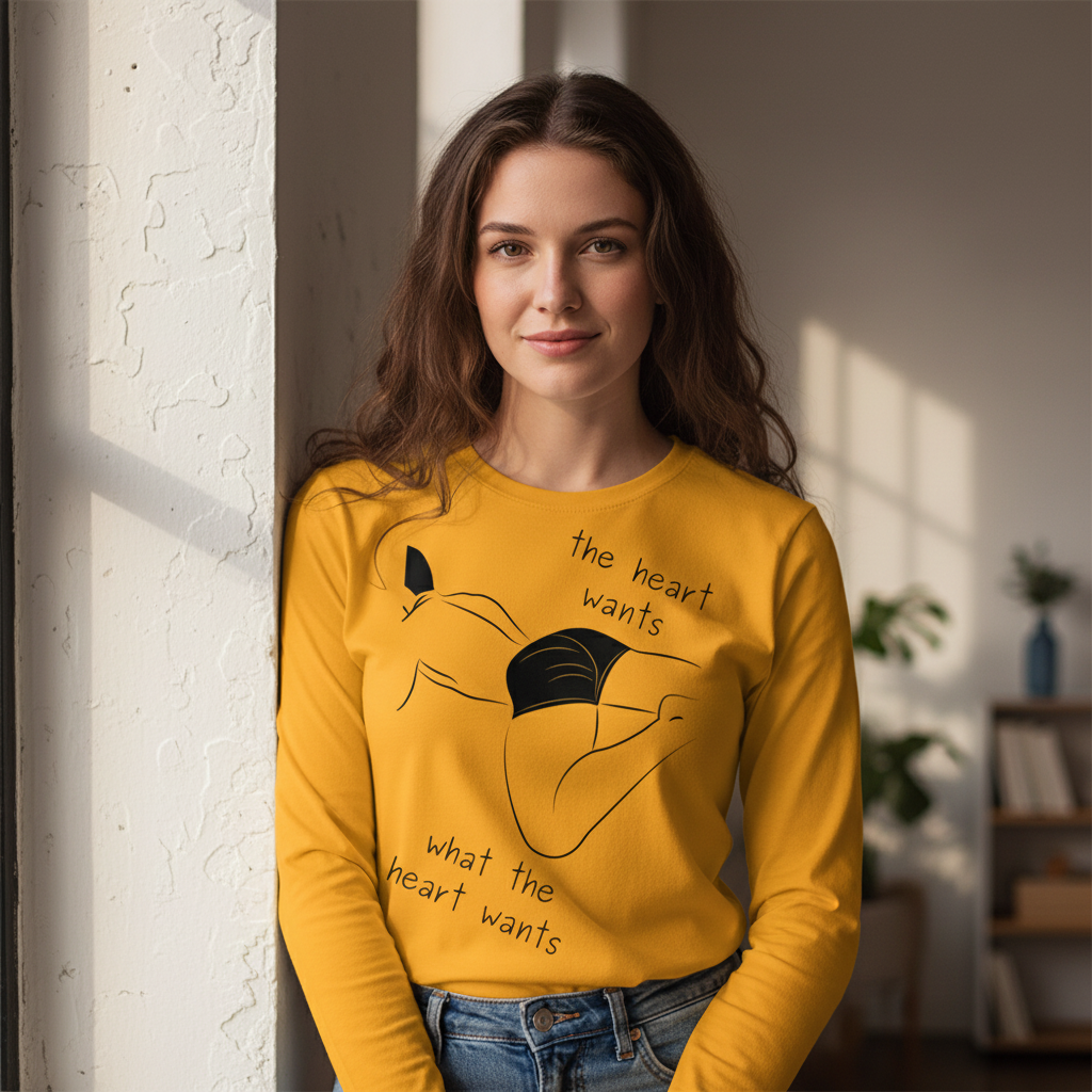 Young woman models The Heart Wants What The Heart Wants Unisex Long Sleeve T-Shirt, featuring a playful illustration and slogan. This mustard-yellow tee celebrates queer pride, identity, and versatile styling.