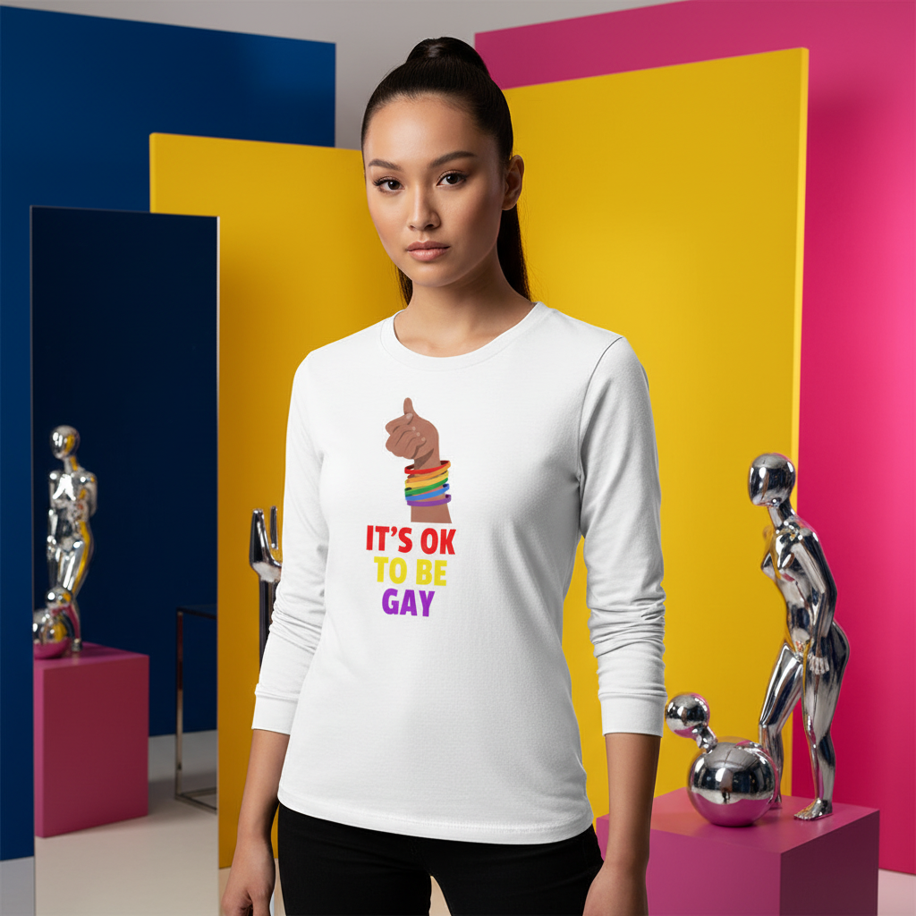 A young person models the It's Ok To Be Gay Unisex Long Sleeve T-Shirt, showcasing a finger-heart print and vibrant slogan, embodying pride and self-expression amidst colorful studio elements.