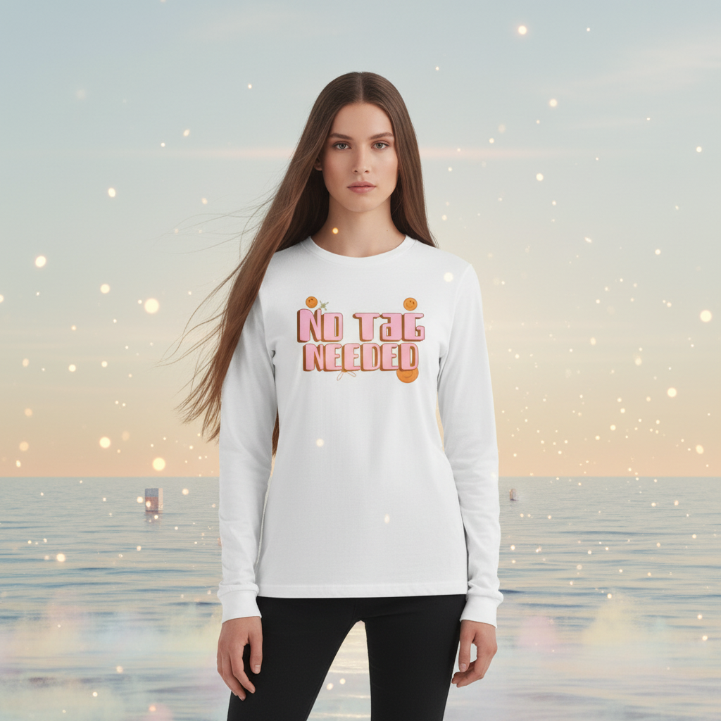 Young woman wears the No Tag Needed Unisex Long Sleeve T-Shirt, featuring pink-and-orange bubble letters with citrus illustrations, set against a dreamy seascape, embodying joyful queer empowerment and self-expression.