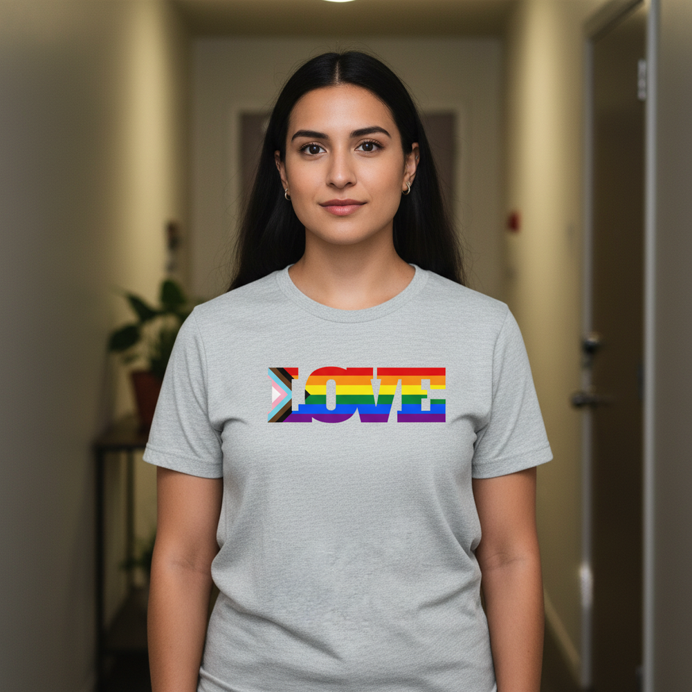 Progress Pride LOVE T-shirt with bold rainbow letters, worn by a woman with long dark hair; hallway and plant in background.