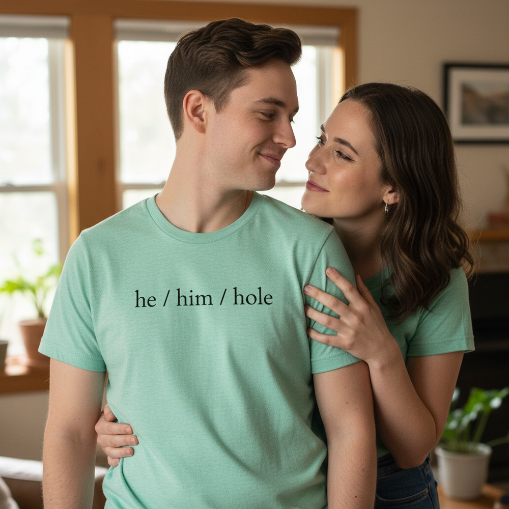 He / Him / Hole T-shirt in light green, modeled by a smiling queer couple indoors; wavy-haired partner hugs from behind.