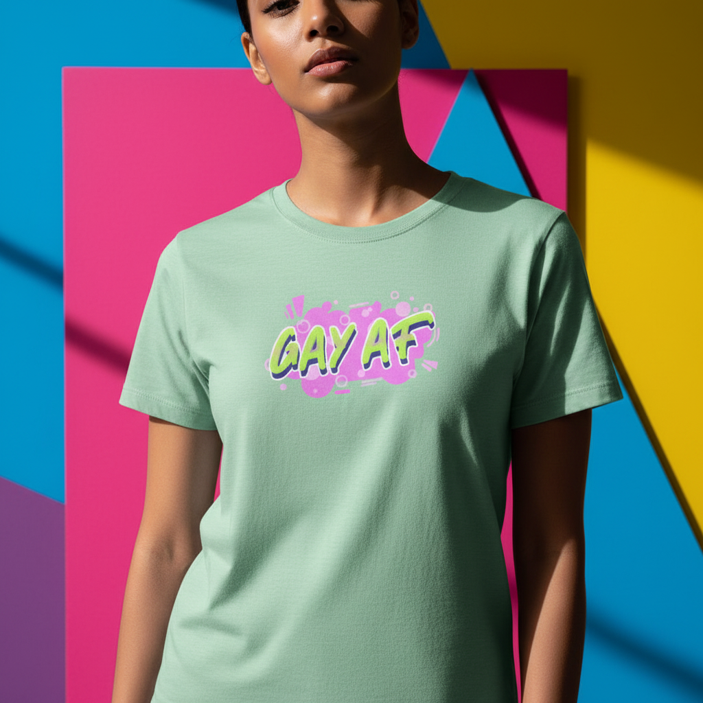 Unisex pale green pride tee with bold, colorful graffiti "GAY AF" text; wearer stands before bright pink, blue, and yellow shapes.