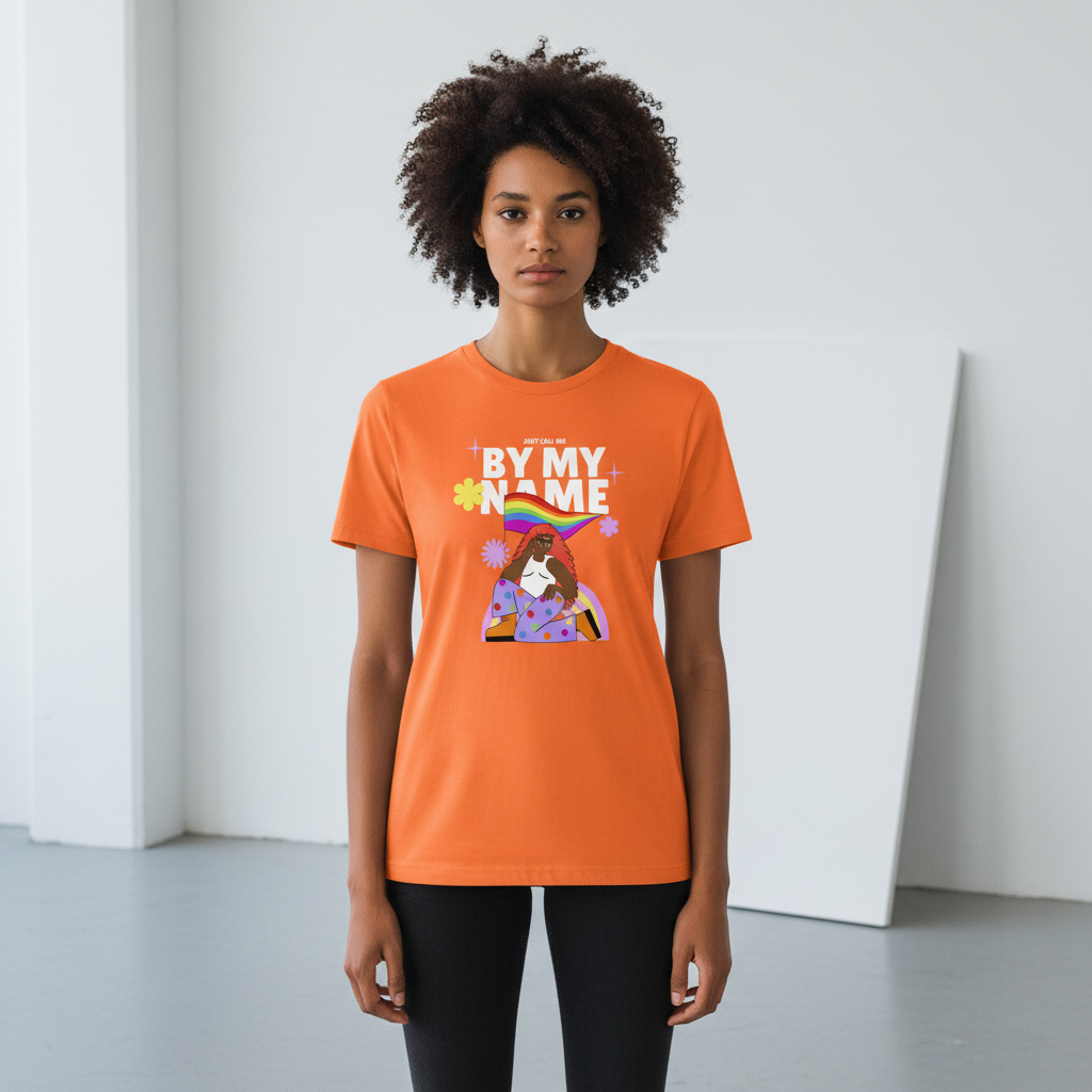 Orange “Just Call Me By My Name” tee with a rainbow Pride flag, florals, and sparkles, worn with black pants on a white backdrop.