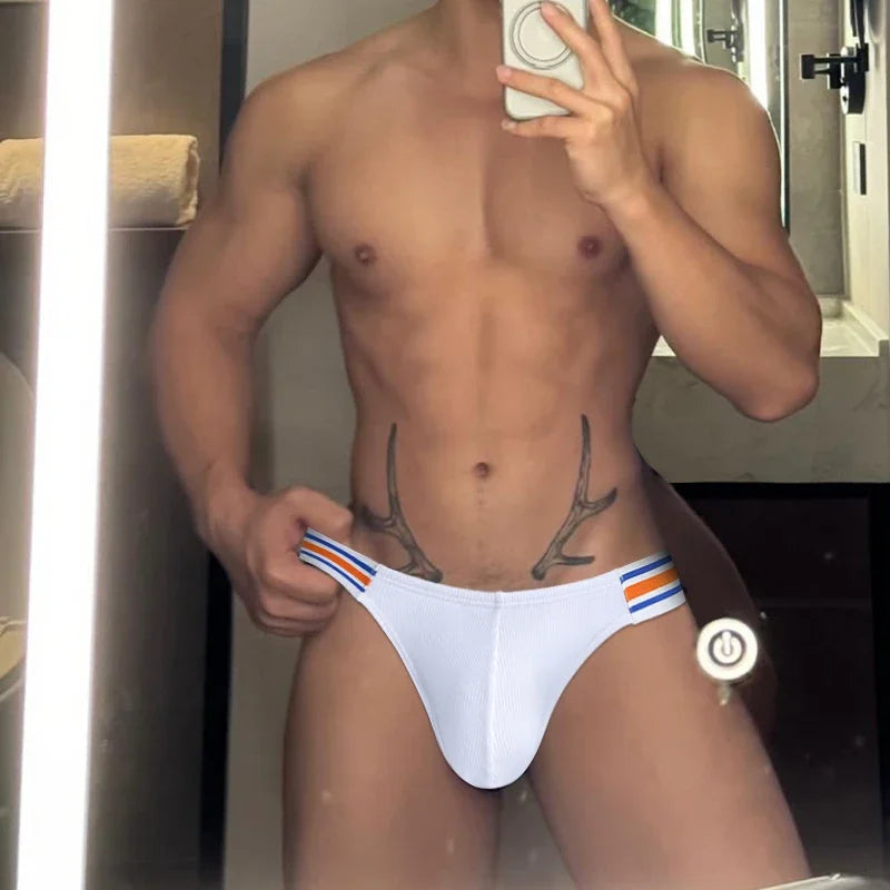 Mirror selfie of a shirtless model wearing white briefs with blue and orange side stripes