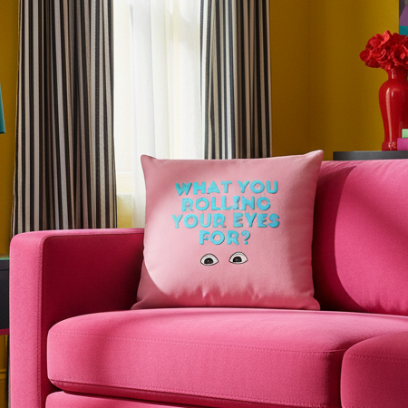 Pink "What You Rolling Your Eyes For?" queer pillow with cartoon eyes and blue text on a bright pink couch
