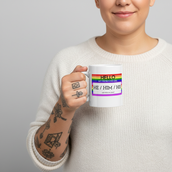 Person holding a white mug with rainbow stripes and “HE / HIM / HIS” pronoun text
