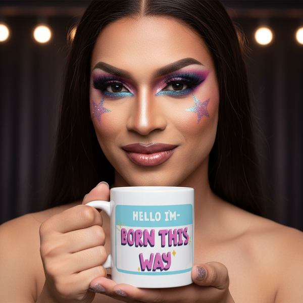 A person with bold purple and teal makeup holds a Hello I'm Born This Way white ceramic mug, embodying vibrant queer pride. The background features softly blurred stage lights, enhancing the empowering, self-expressive vibe.