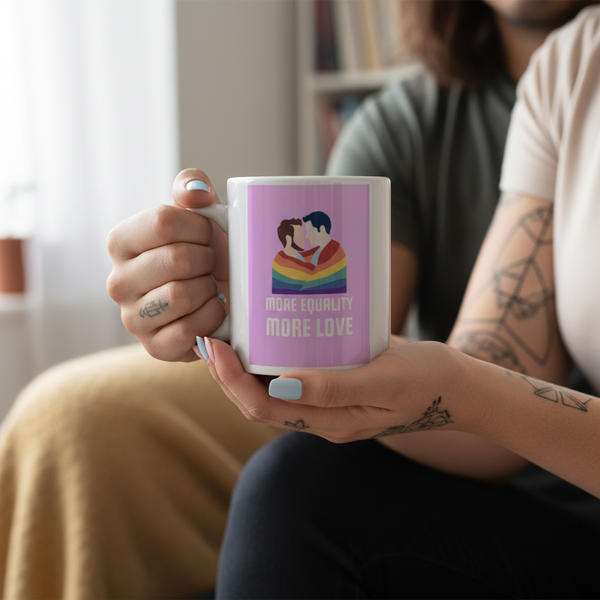 LGBT Couple Mug: two people in a rainbow heart with “MORE EQUALITY MORE LOVE.” Tattooed hand with blue nails holds it; cozy room.