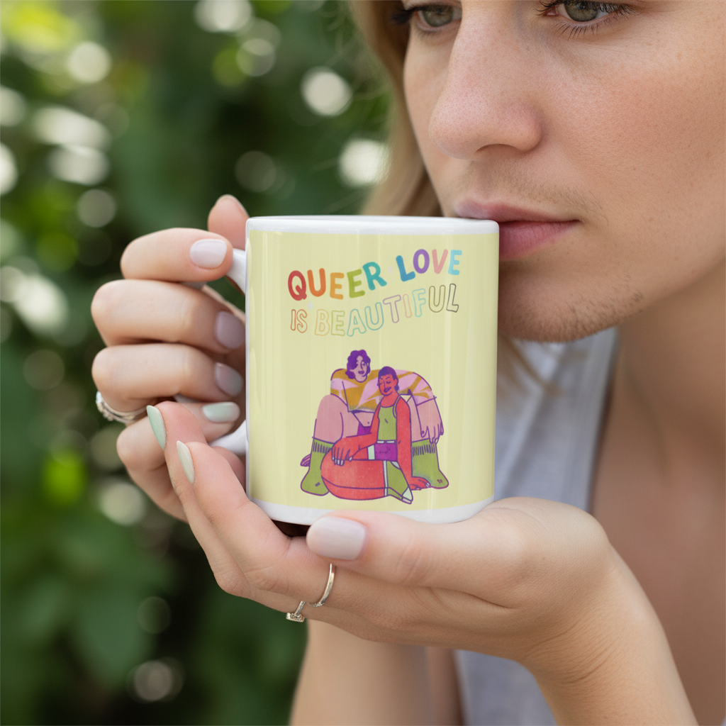 Hands hold a pale yellow mug reading “QUEER LOVE IS BEAUTIFUL” beside a person’s face