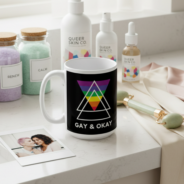 A Gay & Okay mug with rainbow geometric design sits on a vanity with Queer Skin Co. products, bath salts, and a jade roller, embodying vibrant LGBTQIA+ pride and self-care empowerment.