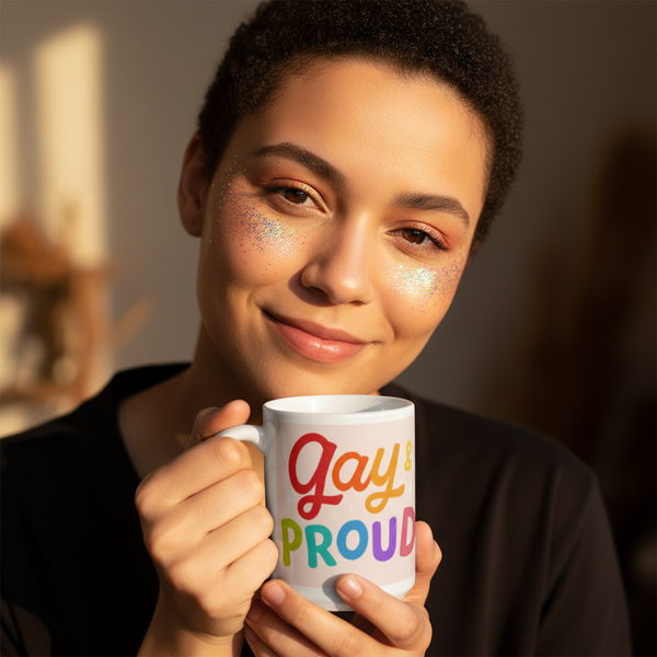 Person with short, curly hair and glitter makeup smiles warmly indoors, holding a Gay & Proud mug. The mug's rainbow letters embody pride, perfect for expressing identity and joyfully celebrating LGBTQIA+ visibility.