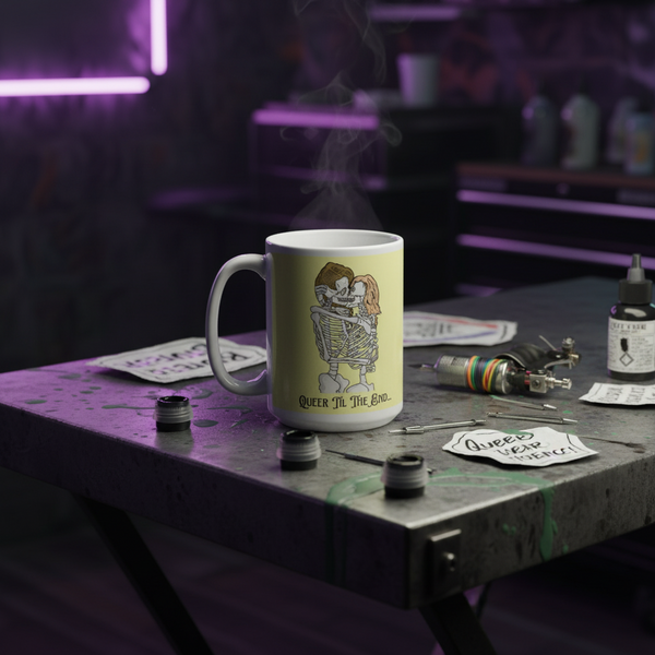 White mug with embracing skeletons and “QUEER ’TIL THE END” text on a tattoo workbench