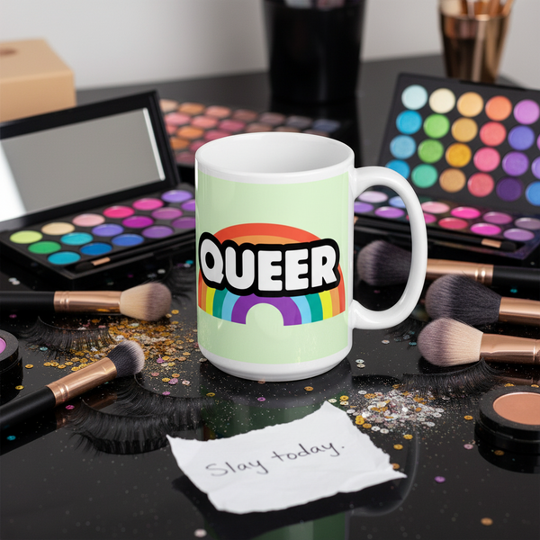 White mug with bold “QUEER” lettering and a rainbow, amid makeup brushes and palettes