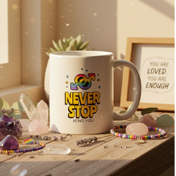 White ceramic Never Stop Being You mug with rainbow gender symbol, surrounded by crystals and beaded bracelets with rainbow charm, embodying pride and self-expression in a sunlit, calming setting.