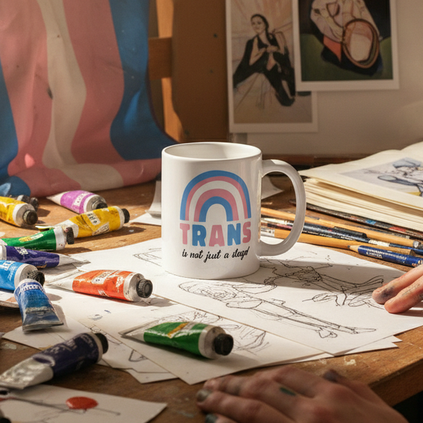 A vibrant artist's workspace features the Trans Is Not Just A Phase! mug, showcasing a rainbow in trans flag colors and empowering slogan, celebrating queer pride and self-expression.
