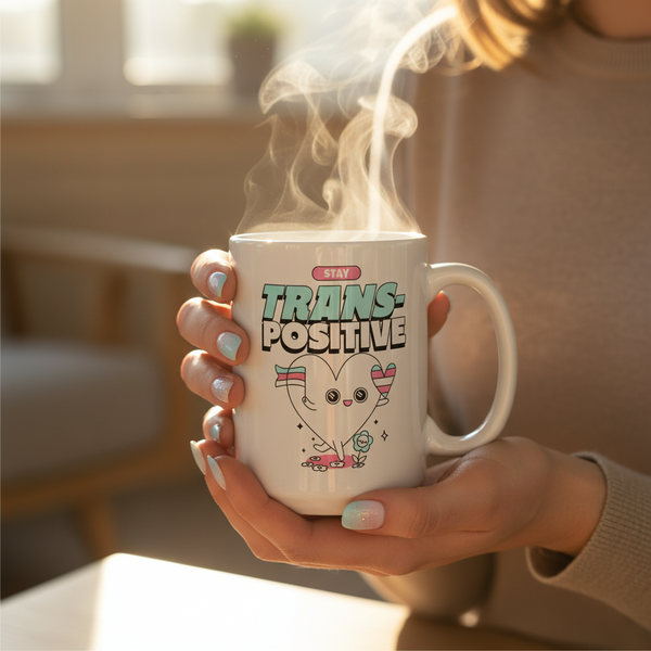 Hands hold the Stay Trans-Positive Mug, featuring a cartoon heart with a trans-flag banner. The sparkly, light-blue nails and steaming mug embody pride and self-expression.
