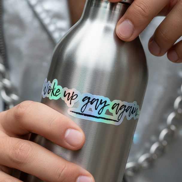 Metallic bottle featuring a bold "Woke Up Gay Again Holographic Sticker." Visible: queer hand, silver chain