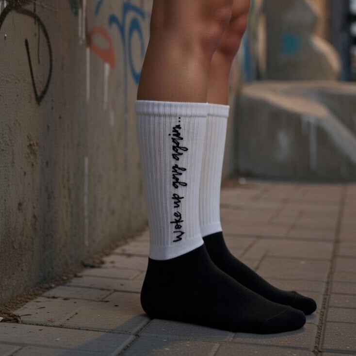 Woke Up Gay Again white crew socks with black cursive "woke up gay again" layered over black socks; graffiti wall behind.