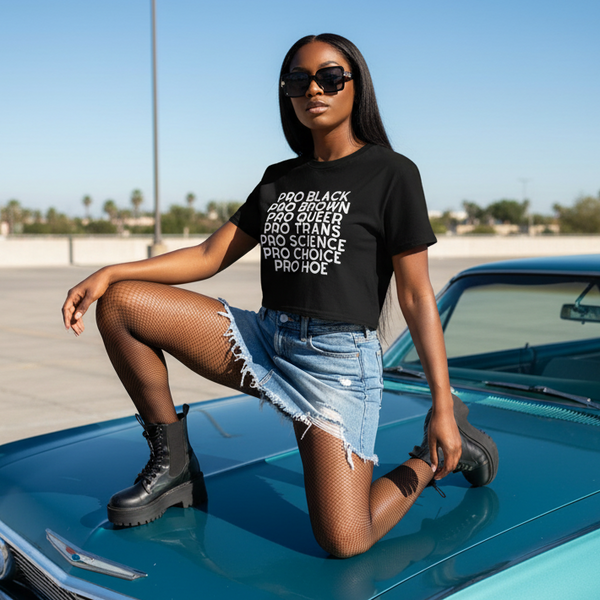 Model kneeling on a teal car wearing a black crop top with stacked “PRO” text and denim shorts