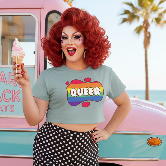 A drag performer wearing the Queer Crop Top, showcasing vibrant self-expression with the QUEER text, paired with polka-dot bottoms, embodying bold pride and joyful queer visibility at a sunlit beach scene.