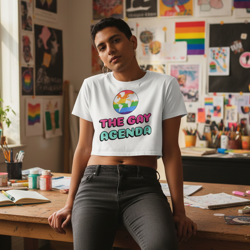 Person wearing The Gay Agenda Crop Top, featuring a rainbow-striped globe and bold text, casually leans against an art-filled studio table covered with creative supplies, embodying queer pride and self-expression.