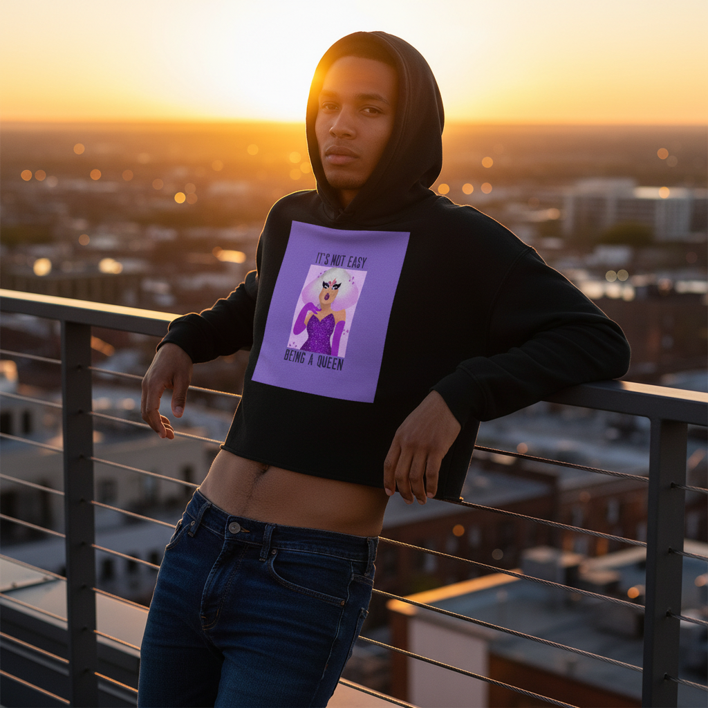 It's Not Easy Being A Queen" crop hoodie with cartoon graphic, worn by a young man in jeans on a sunset-lit city rooftop.