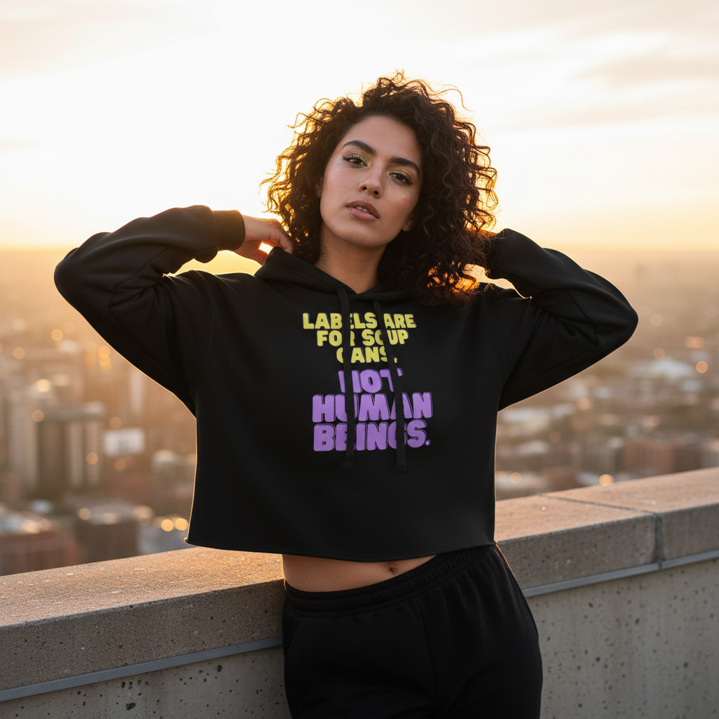 Crop hoodie with bold yellow and purple text, “Labels are for soup cans. Not human beings.” Queer woman poses at sunset rooftop.