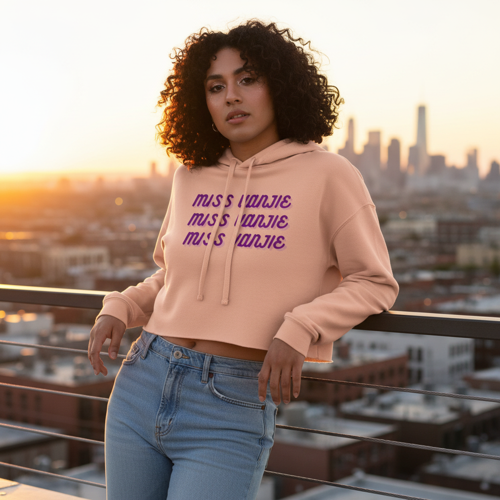 Miss Vanjie Crop Hoodie in light pink with “MUSIQUE” in bold purple, rocked by a curly-haired woman on a city rooftop at sunset; paired with light blue jeans for a standout queer moment.