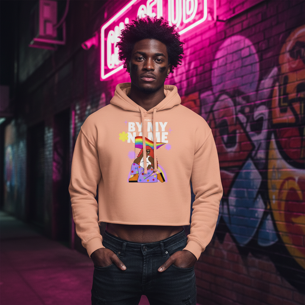Peach "Just Call Me By My Name" crop hoodie with Pride rainbow flag, modeled by a young Black man in front of neon-lit graffiti walls.