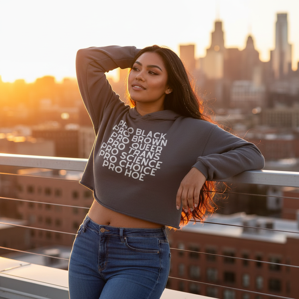 Gray Pro Hoe Crop Hoodie with bold white “pride” text, worn by a woman on a rooftop at sunset, city skyline glowing.