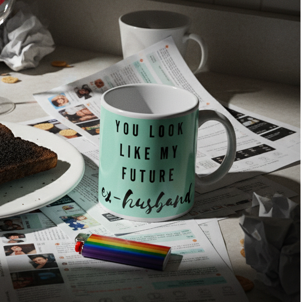 Mint-green “You Look Like My Future Ex-husband” mug on newspapers beside a rainbow lighter and toast