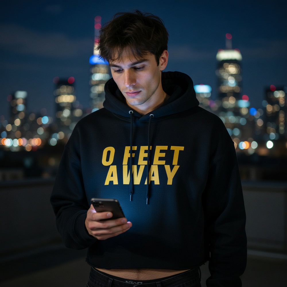 Zero Feet Away Grindr crop hoodie with bold yellow “0 FEET AWAY” text, worn by a young man at night in the city.
