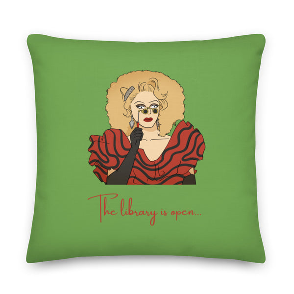 Green square pillow with a drag performer illustration and red text reading “The library is open...”