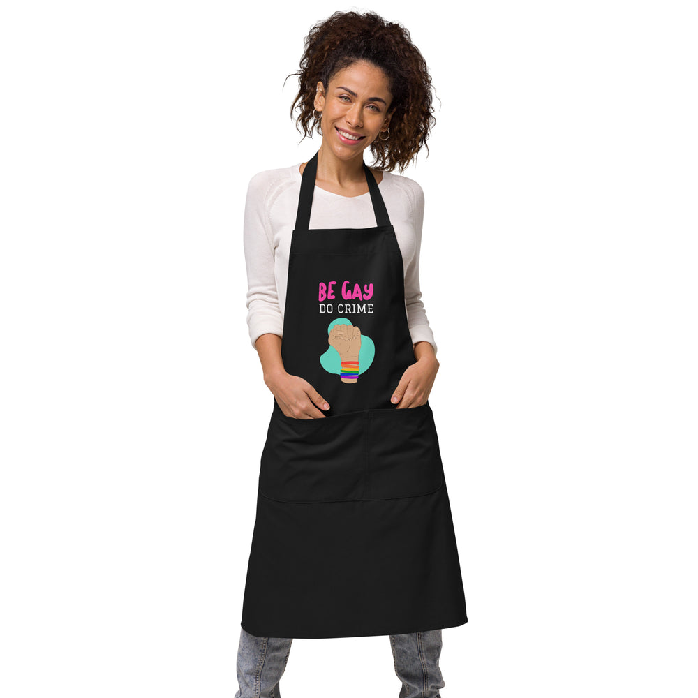 Model wearing a black apron reading “BE GAY DO CRIME” with a raised fist and rainbow wristbands