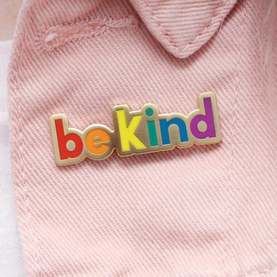 Close-up of multicolored “be kind” enamel pin on pink fabric