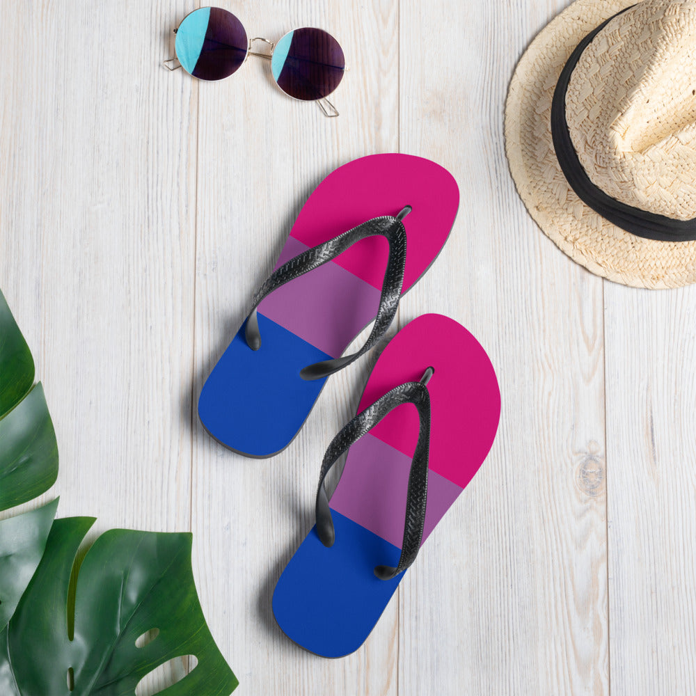 Bisexual Pride flip-flops with pink, purple, and blue stripes on gray, styled with pink ribbon, beads, amethyst, pearl, and dish.