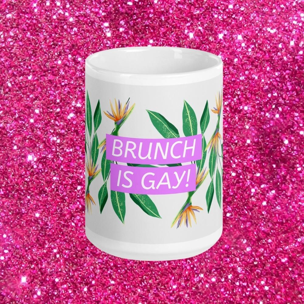 White mug reading “BRUNCH IS GAY!” with tropical flowers against a pink glitter background