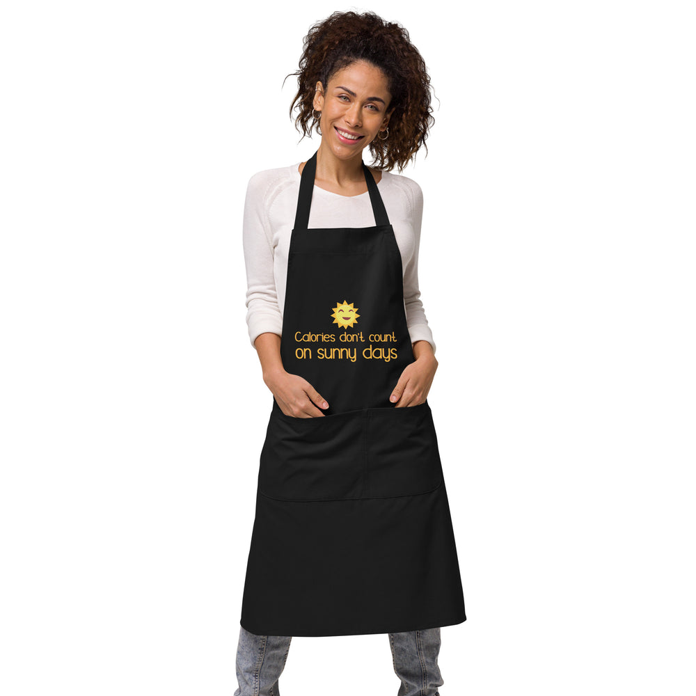 Front view of a model wearing a black apron with a smiling sun and “Calories don't count on sunny days”