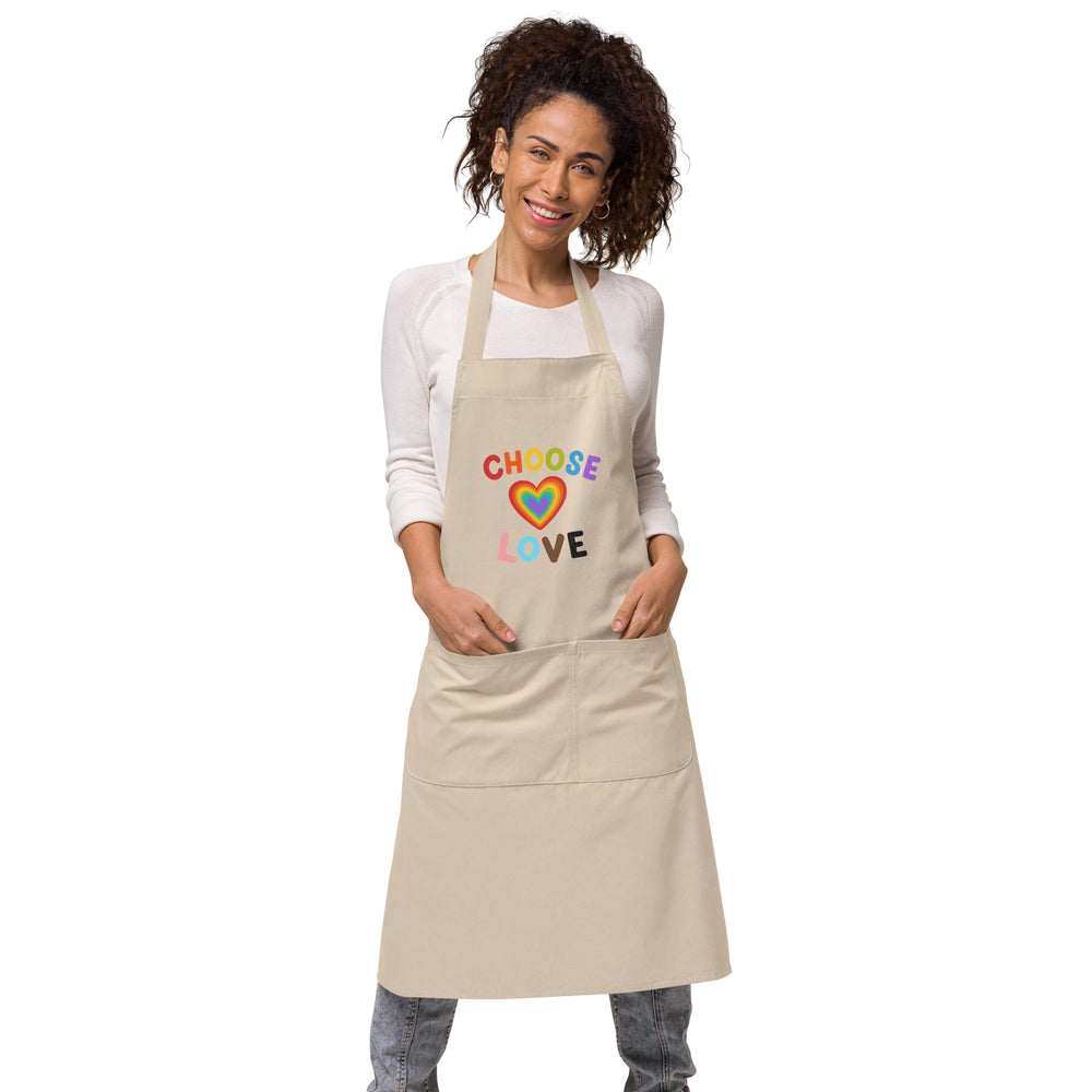 Front view of a beige apron modeled by a smiling person, reading “CHOOSE LOVE” above a rainbow heart