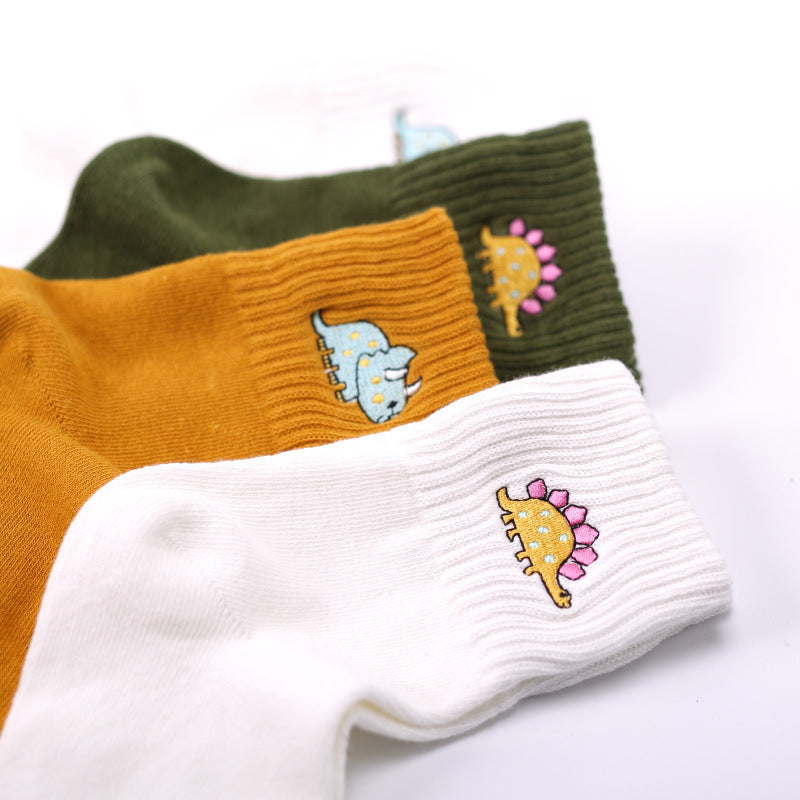 Three ribbed dinosaur socks in mustard, olive green, and white with colorful dinosaur embroidery