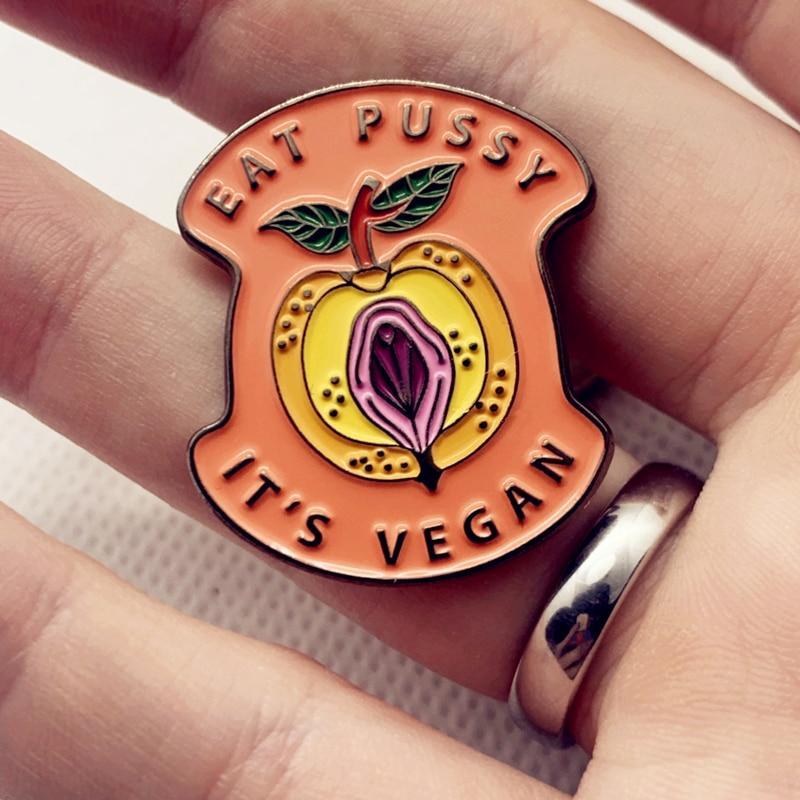 Close-up of an enamel pin reading “EAT PUSSY IT’S VEGAN,” with a peach and fruit design