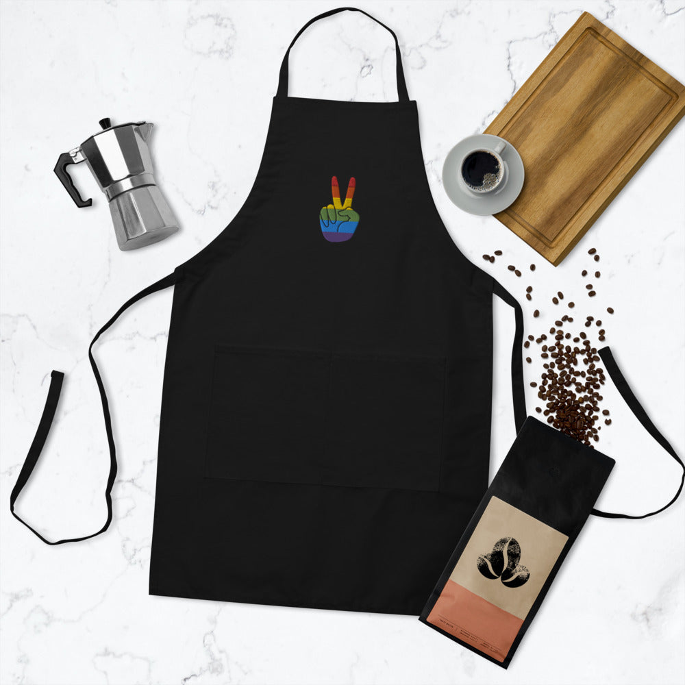 Black apron laid flat with a rainbow peace-sign hand emblem, pocket, and waist ties