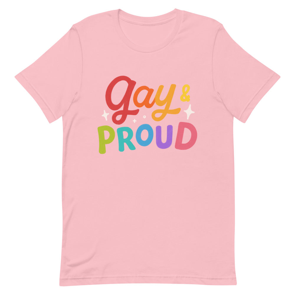 Pink “Gay & Proud” T-shirt and denim jacket worn by queer women hugging at sunny Pride, rainbow flags and joyful crowd behind.