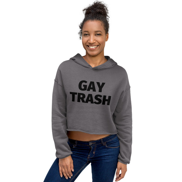 Crop hoodie with bold “Gay Trash” black text, worn by a young man in blue jeans on a rooftop at sunset, city skyline behind him.
