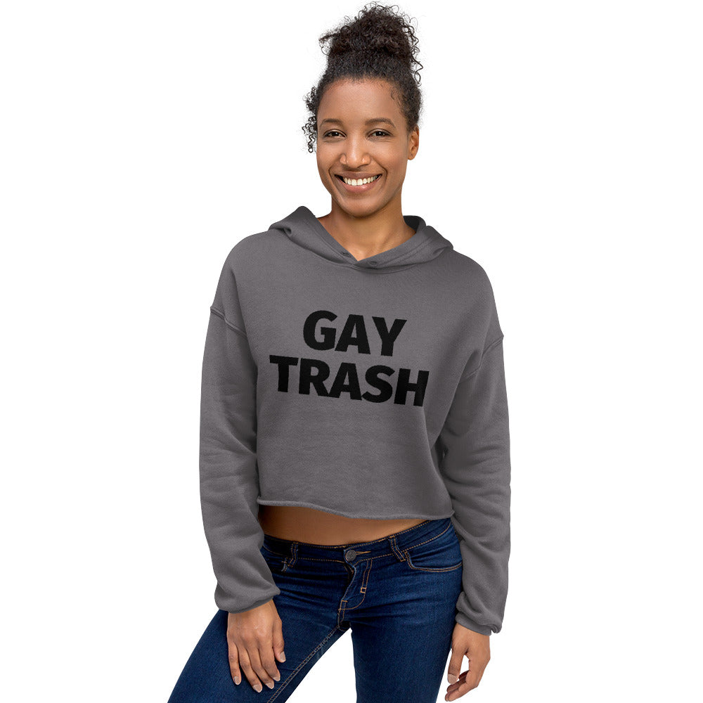 Crop hoodie with bold “Gay Trash” black text, worn by a young man in blue jeans on a rooftop at sunset, city skyline behind him.