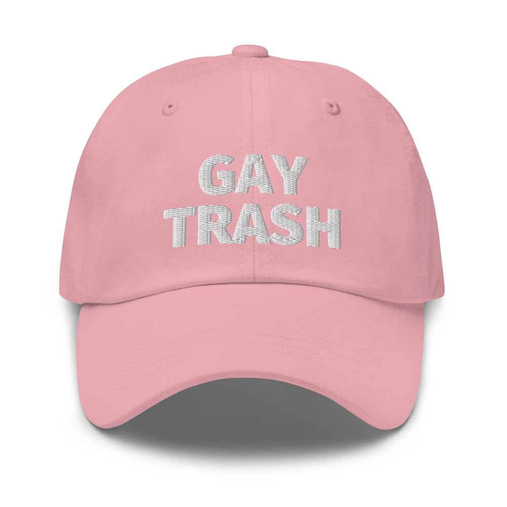 Light pink embroidered dad hat reads "GAY TRASH" in bold white letters; queer person with short curls, hoop earrings, smiling.