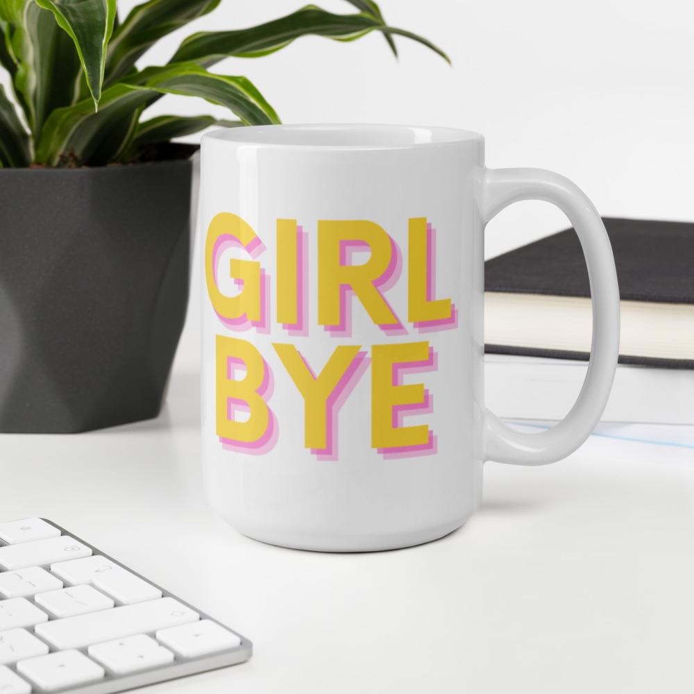White mug reading “GIRL BYE” in yellow and pink lettering beside a plant and keyboard